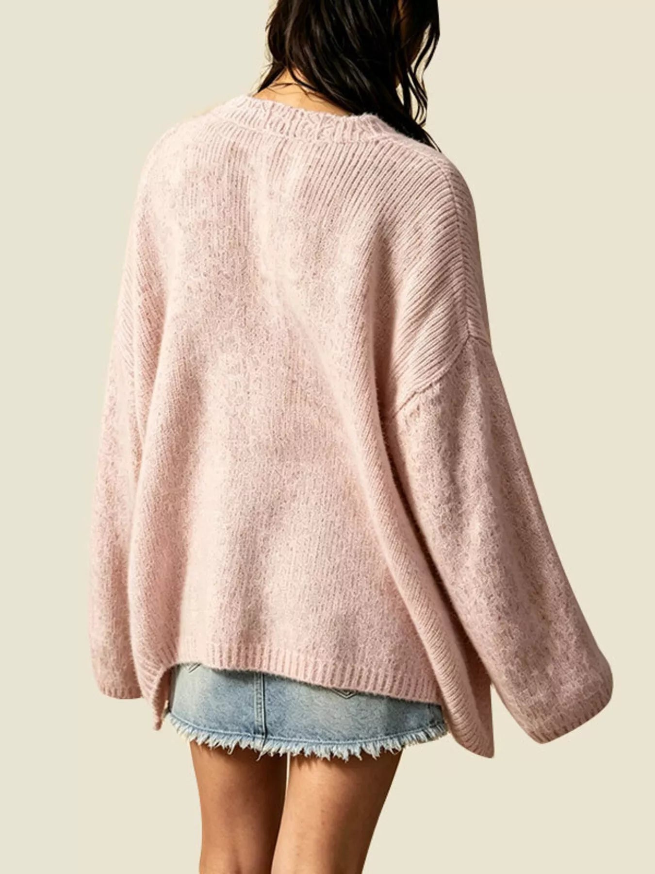 A person shown from the back wearing a loose, oversized pink knit sweater with long sleeves and a ribbed crew neckline, paired with light blue denim shorts against a plain light beige background.