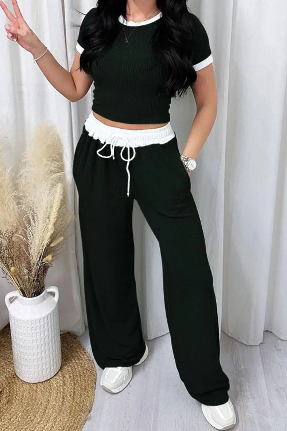 Person wearing a black and white outfit with wide-leg pants and a crop top, standing against a white curtain.