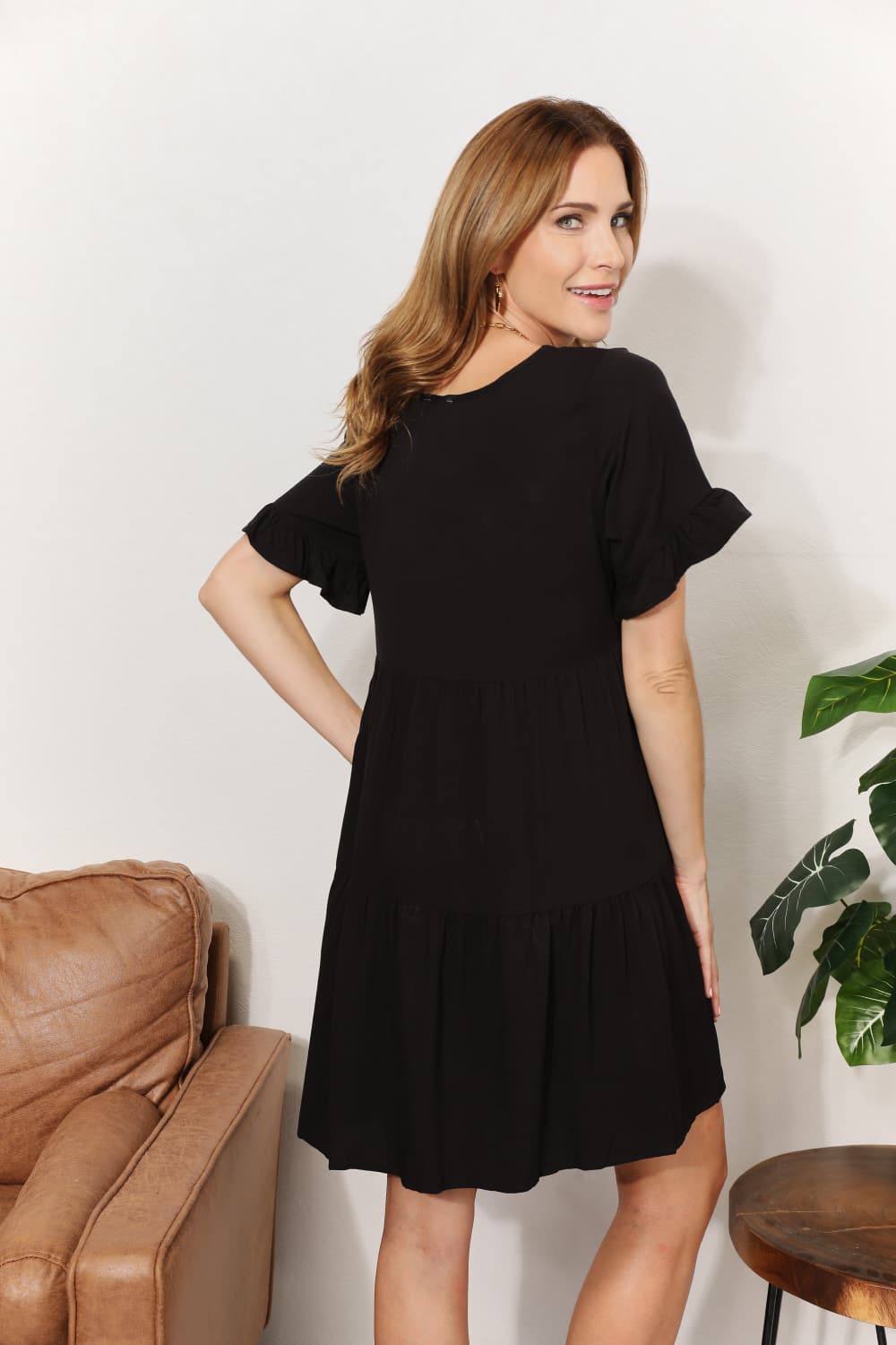 Woman wearing a black dress standing in a room with a brown leather couch and a plant.