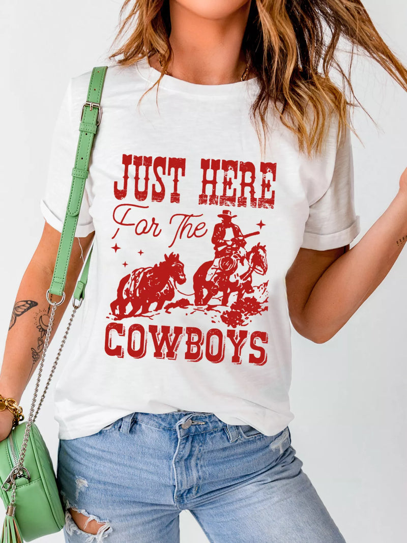 A woman wearing a white graphic t-shirt with a red 'Just Here For The Cowboys' print, paired with distressed blue jeans and a green shoulder bag.