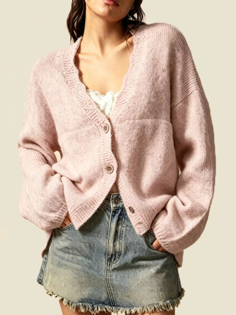A woman wearing a light pink fuzzy knit cardigan with a V-neck and button closure, paired with a white top and denim skirt, against a solid beige background.