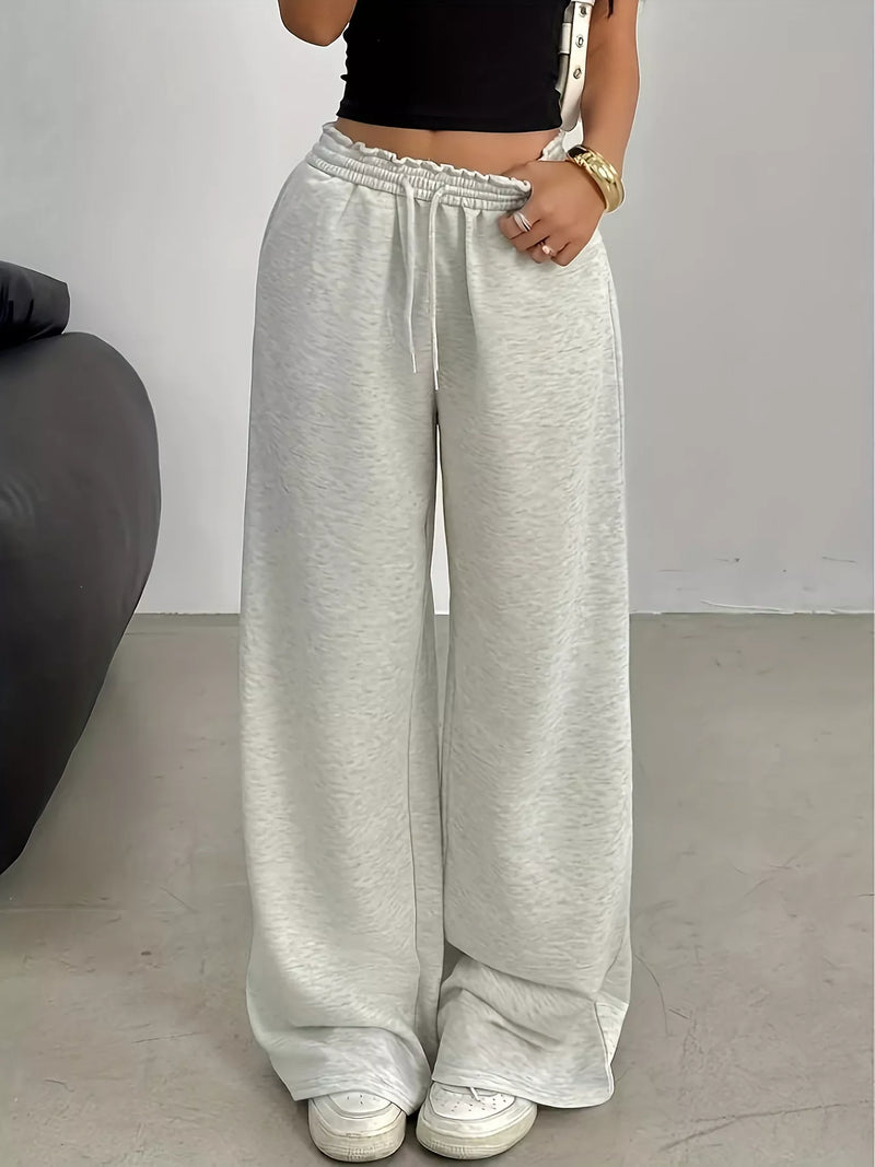 A person wearing light heather gray wide-leg sweatpants with an elastic drawstring waist, paired with a black crop top and white sneakers, standing indoors against a plain light gray wall.