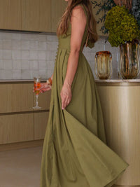 Woman in a green dress standing in a kitchen with a vase of flowers and a drink in the background.