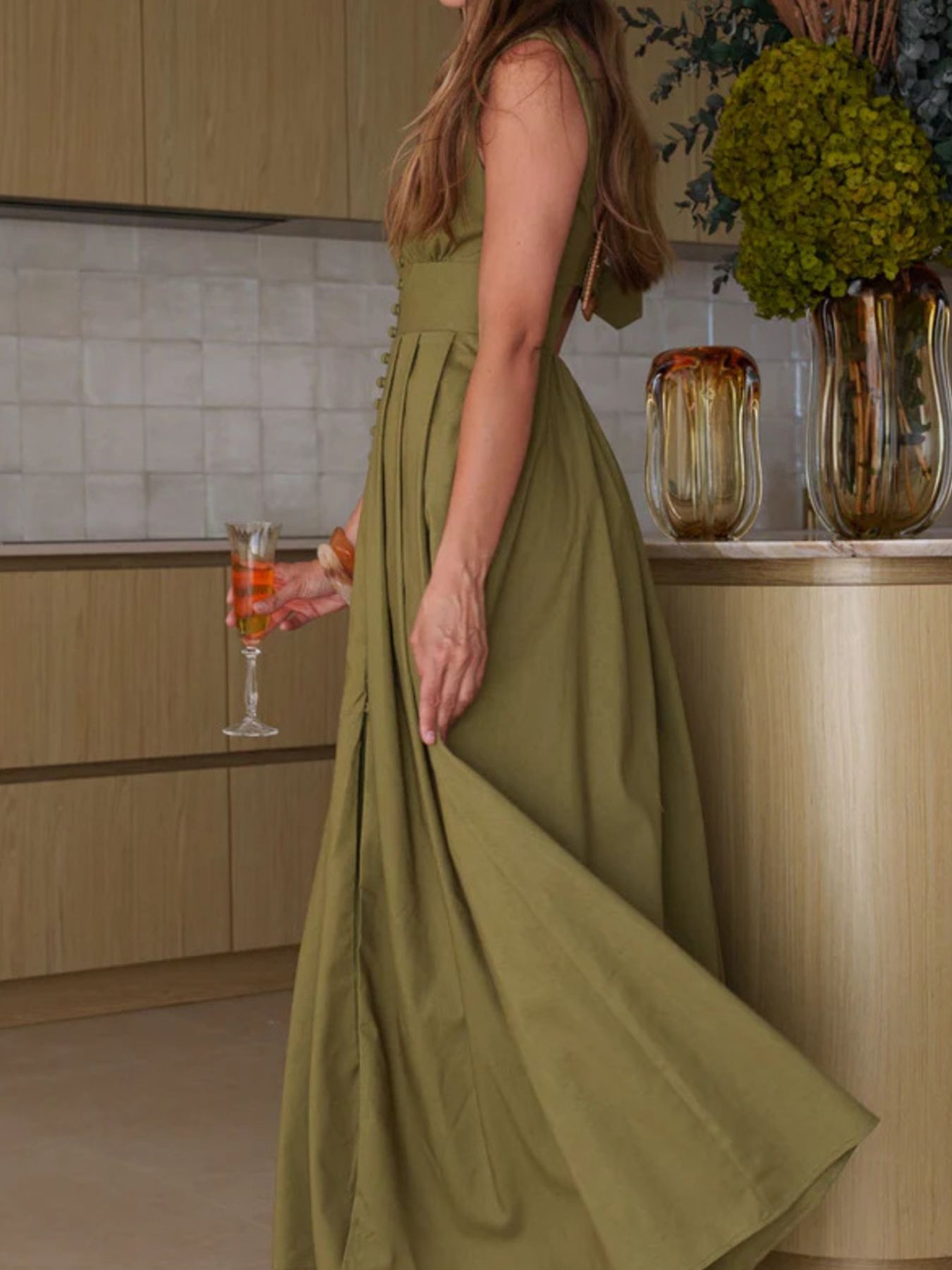 Woman in a green dress standing in a kitchen with a vase of flowers and a drink in the background.