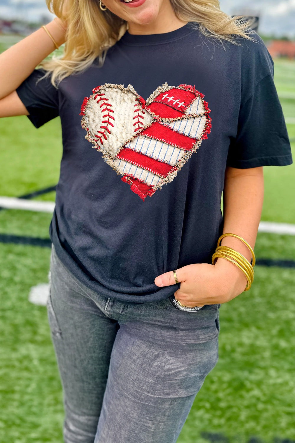 Person wearing a navy t-shirt with a heart design featuring a baseball and American flag on a sports field.