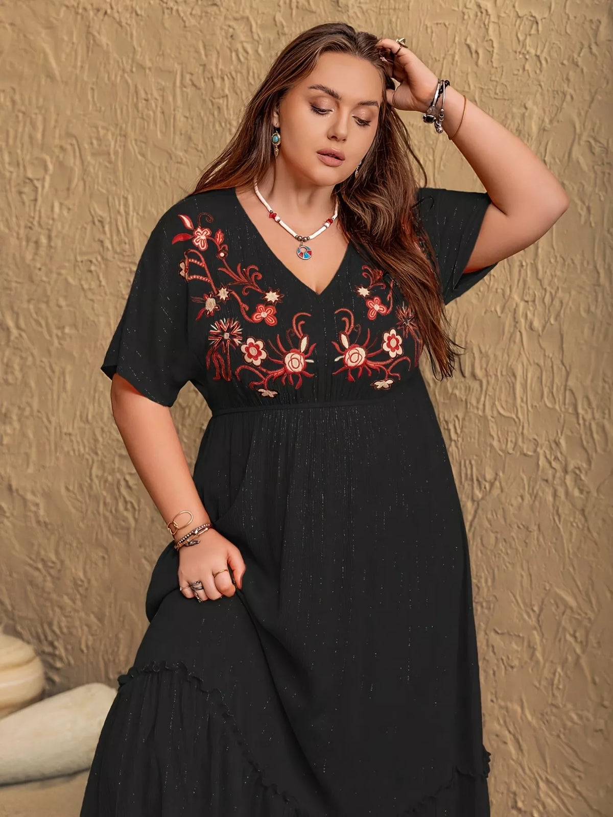 Woman wearing a black dress with red floral embroidery against a textured beige wall.
