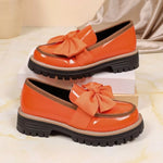 A pair of women's chunky platform loafers in glossy orange patent leather, each featuring a matching orange fabric bow on the instep and a thick black lug sole, displayed on white and beige boxes against a light marble surface.