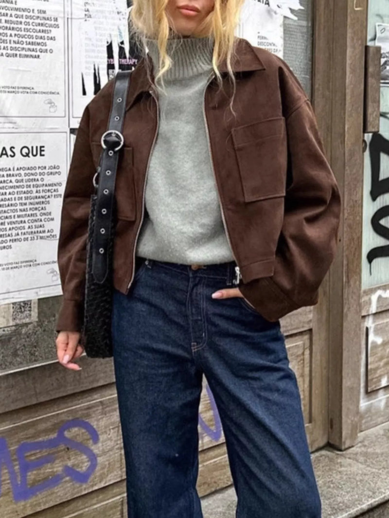 A woman wearing a brown suede-like utility jacket over a gray turtleneck sweater and dark blue jeans, standing against a city wall with posters and graffiti.
