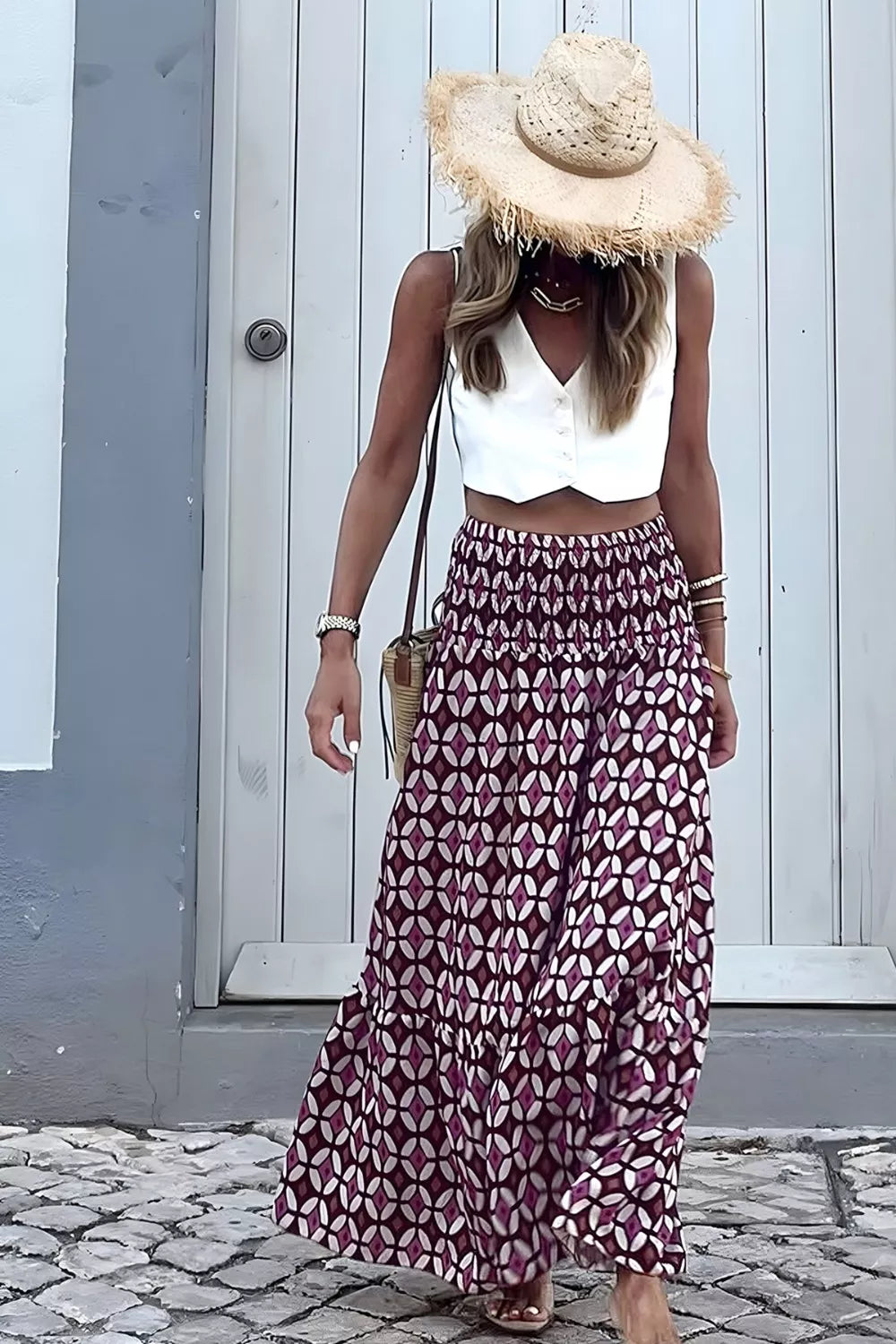 Woman wearing a white top and patterned skirt with a straw hat, standing on a cobblestone street.