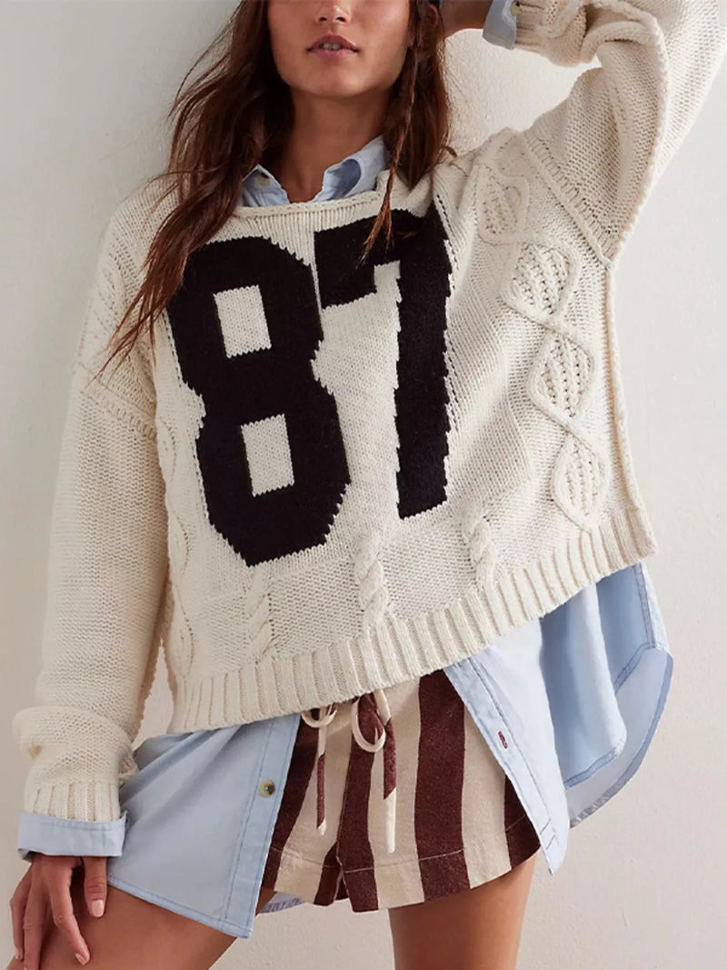 A woman wearing a cream cable-knit sweater with a large black '87' graphic on the front, layered over a light blue button-up shirt and brown-and-white striped shorts.