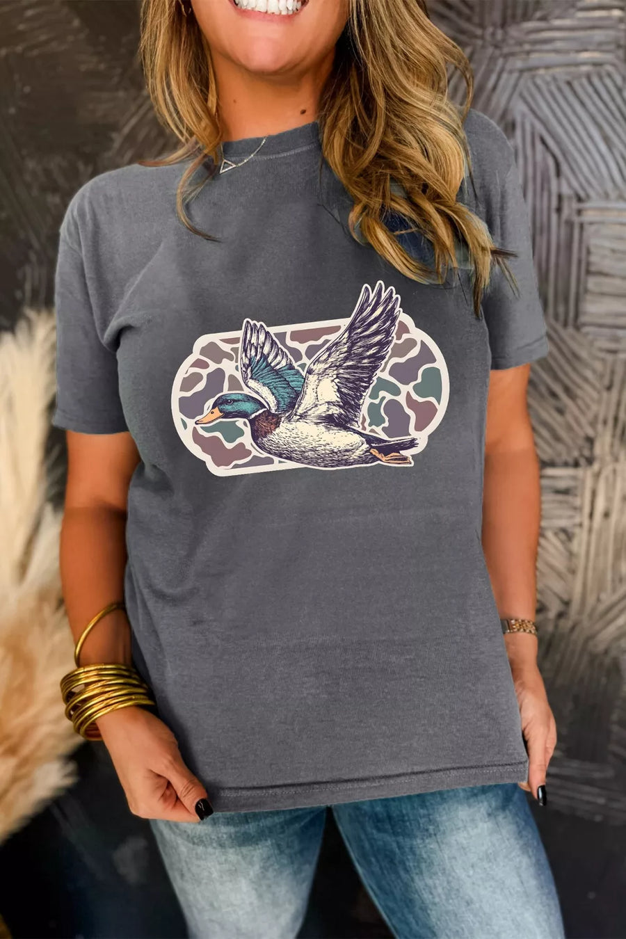 Person wearing a gray t-shirt with a colorful graphic of a bird, standing against a textured wall.