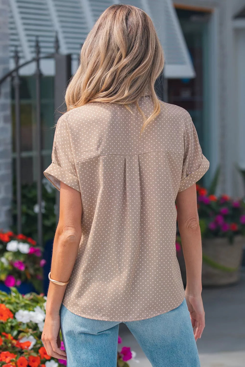 Woman wearing a beige patterned shirt and blue jeans standing in front of a building with plants.