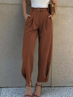 A person wearing high-waisted, wide-leg brown pants with a matching belt, styled with a white top and brown strappy sandals against a textured beige wall.