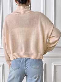 Person wearing a beige sweater and blue jeans against a white paneled wall.