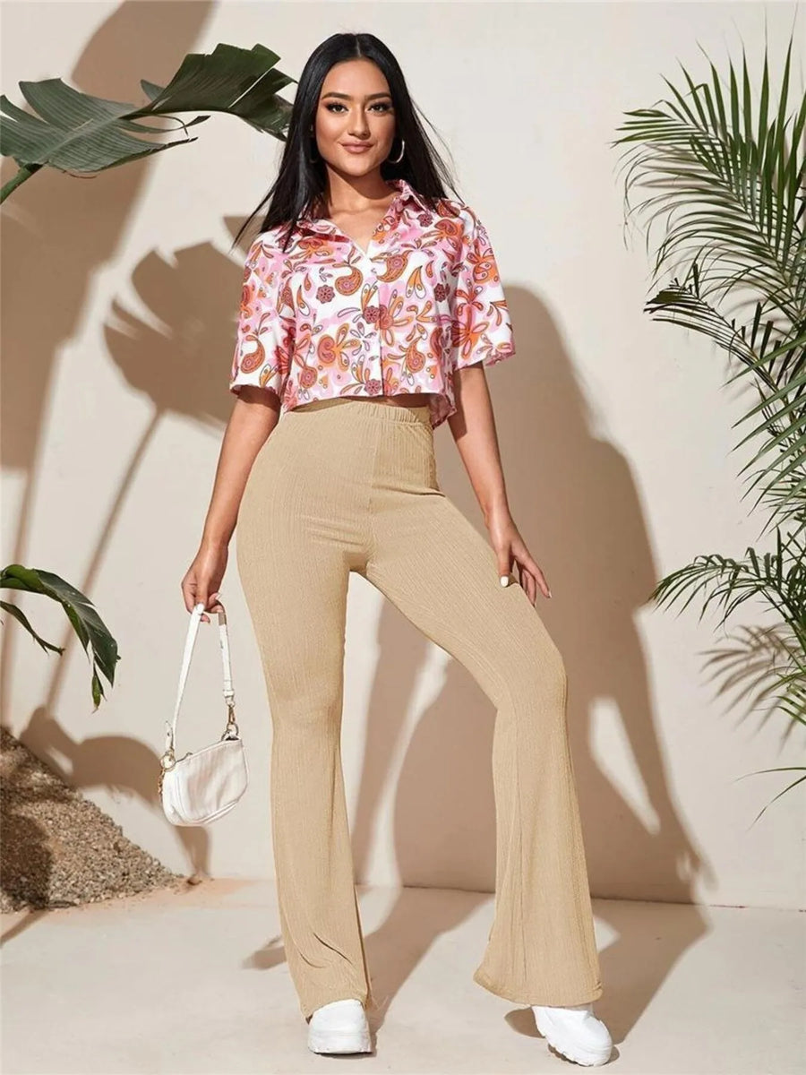 Woman wearing a floral blouse and beige pants standing against a neutral background with plants.