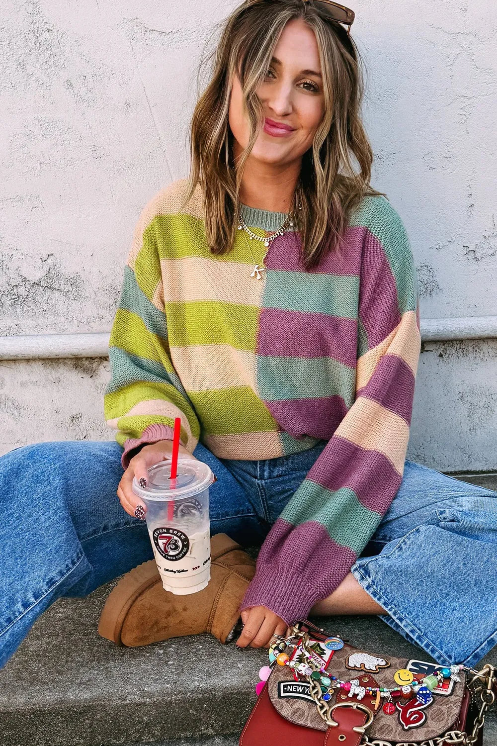 Woman wearing a colorful striped sweater sitting on steps with a drink and small bag.