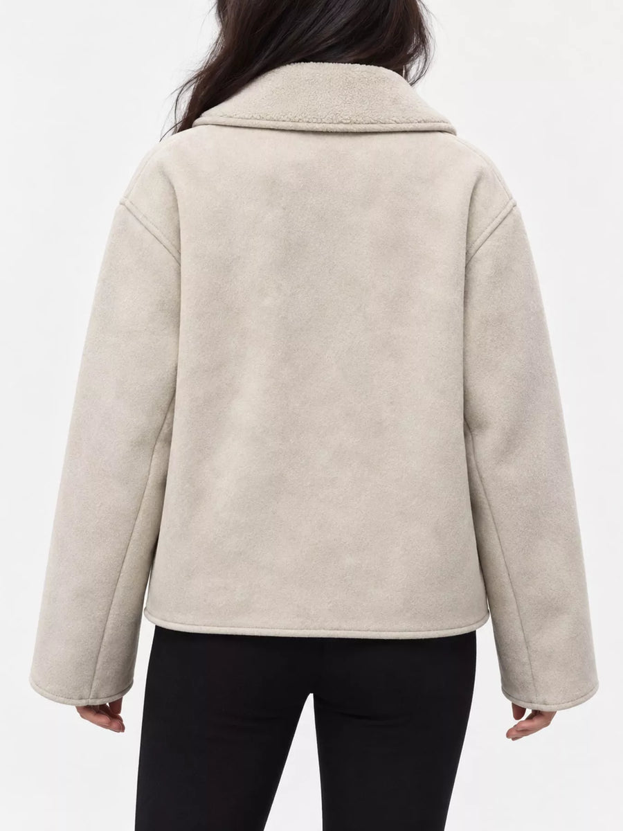 A person shown from the back wearing a light beige sherpa or fleece jacket with a wide collar and long sleeves, paired with black pants, against a plain white studio background.