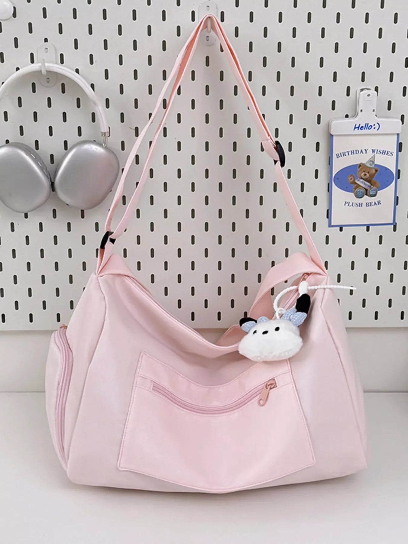 Pink handbag with a small plush toy attached, on a white surface with a pegboard background.