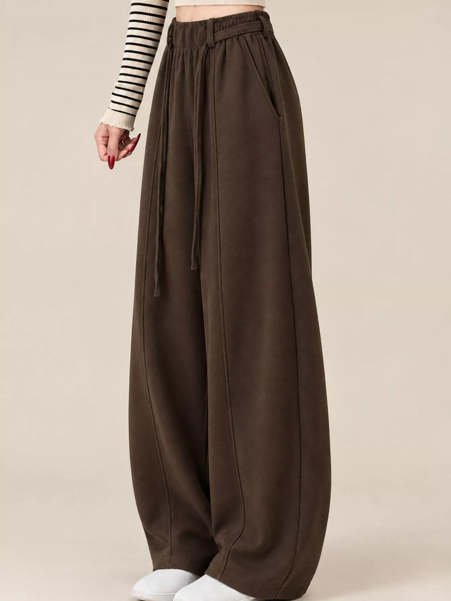 A person wearing very wide-leg, high-waisted brown pants with a drawstring waistband, paired with a black-and-white striped long-sleeve top and white slip-on shoes, against a plain beige studio background.