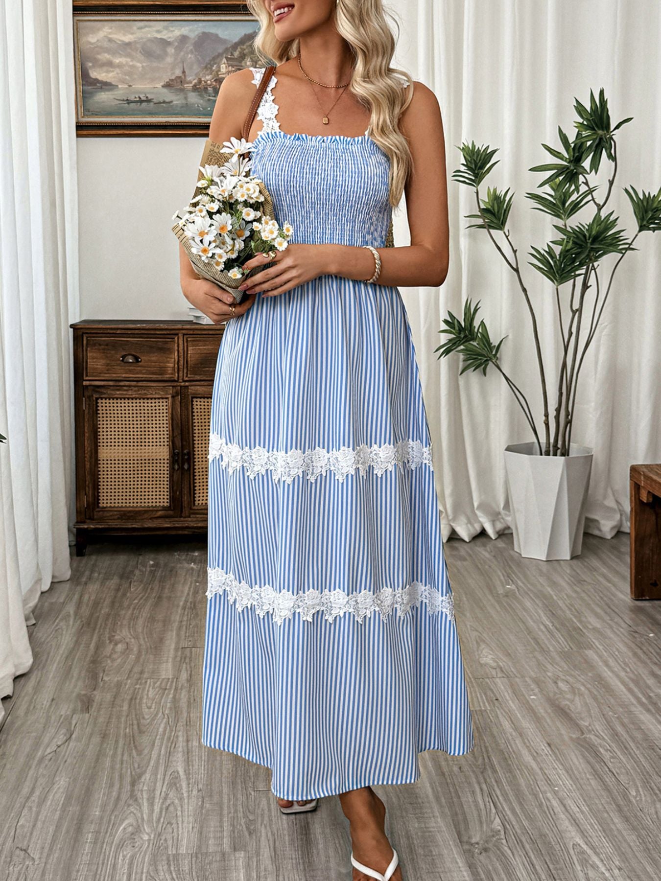 Woman in a blue striped dress holding flowers in a room with plants and furniture.