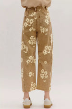 A person shown from the waist down wearing tan wide-leg cropped jeans with a cream floral pattern, paired with white sneakers, against a light gray studio background.