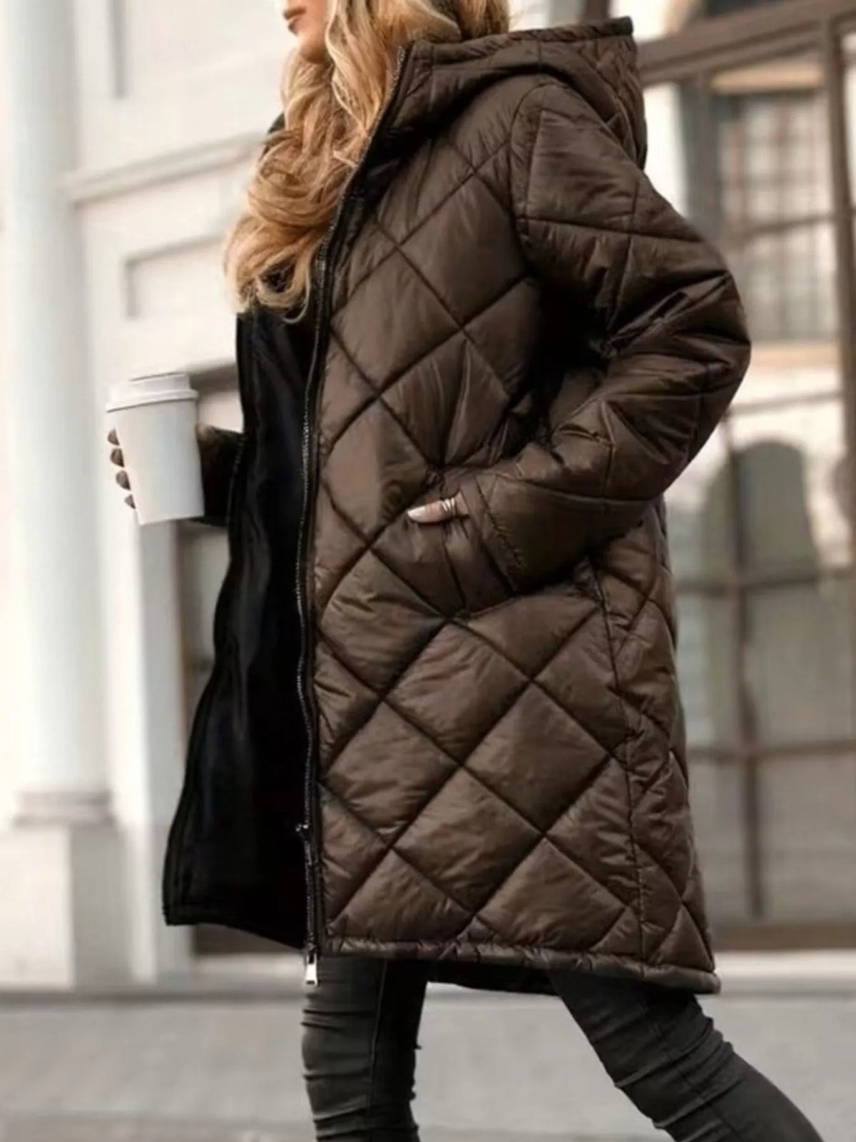 A woman wearing a long quilted brown puffer coat with a hood, walking outdoors while holding a white coffee cup.
