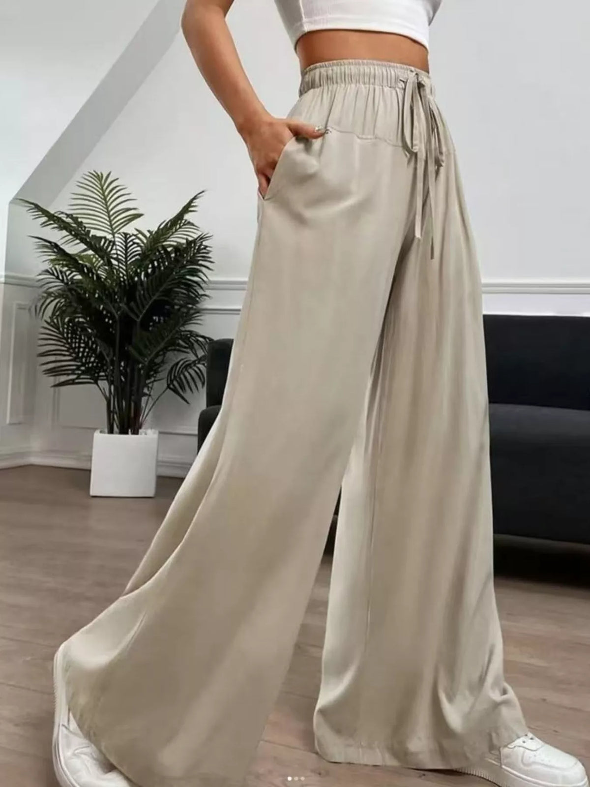 A person wearing loose, wide-leg beige pants with an elastic drawstring waist, paired with a white crop top and white sneakers in a modern indoor room with a plant and dark sofa.