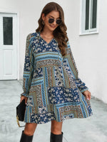 Woman wearing a blue patterned dress with black boots and sunglasses, standing in front of a white wall.