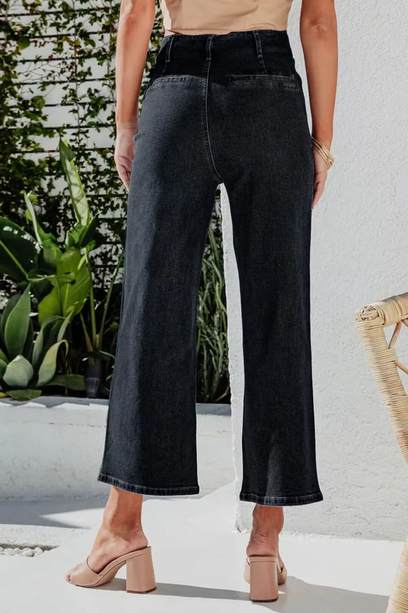 Person wearing black wide-leg jeans standing outdoors with plants and a white wall in the background.