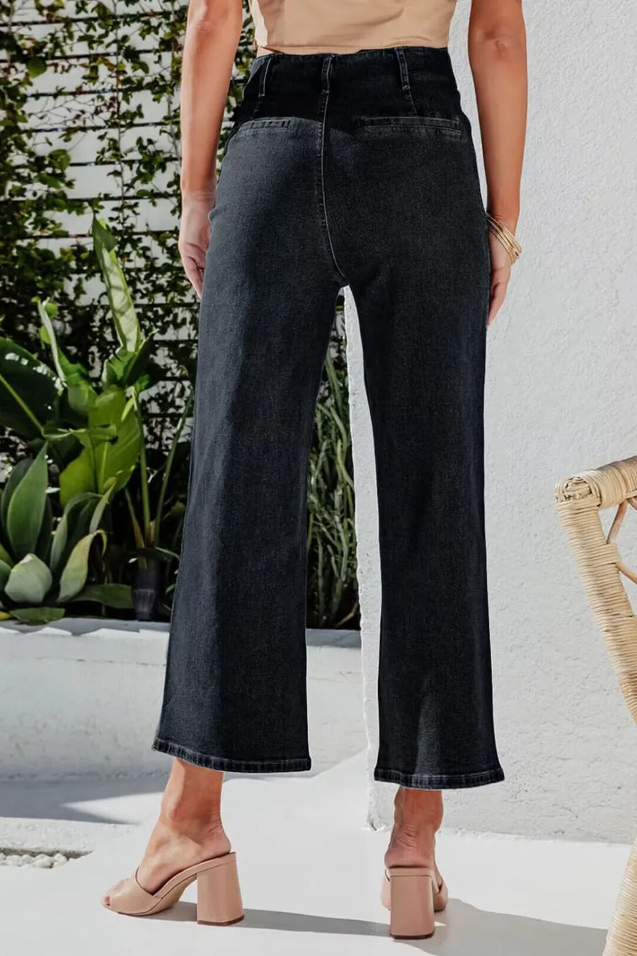 Person wearing black wide-leg jeans standing outdoors with plants and a white wall in the background.