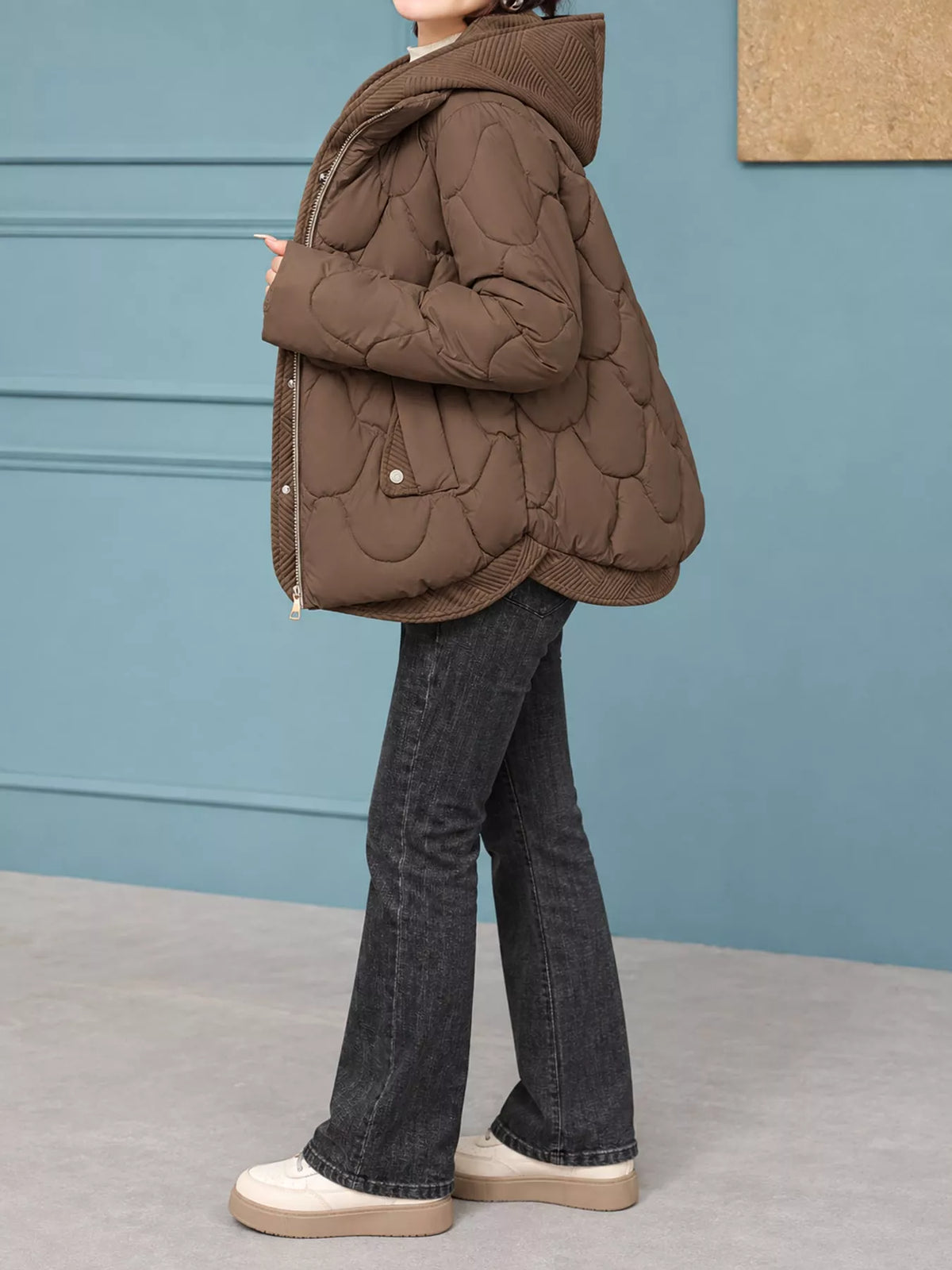 A person wearing a brown quilted puffer jacket with a hood, paired with dark gray flared jeans and white platform sneakers, standing against a blue paneled wall.