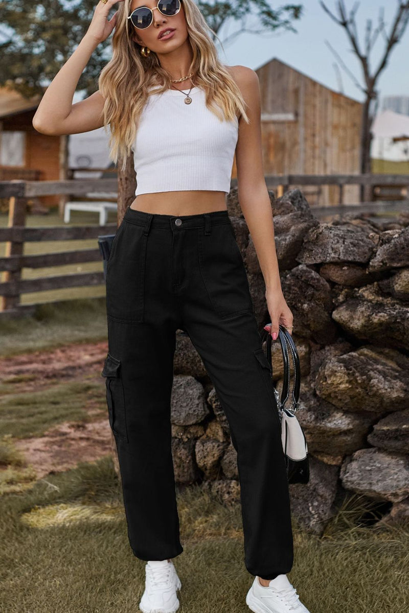 Woman in a white crop top and black cargo pants standing outdoors near a stone wall.