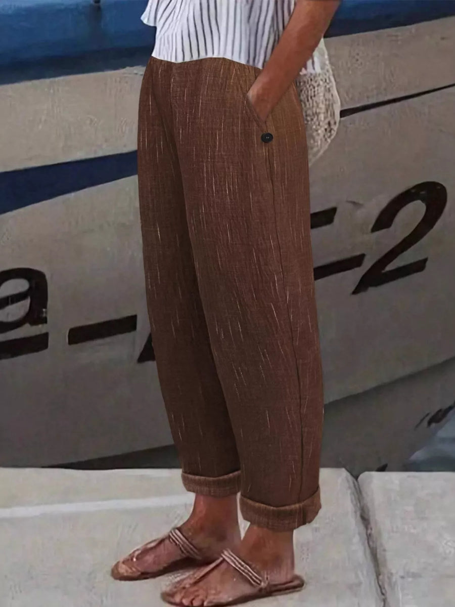 A person wearing loose brown textured cropped pants with rolled cuffs, paired with a white striped top and flat sandals, standing outdoors in front of a boat.