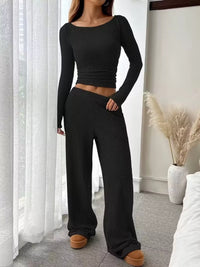 Person wearing a black ribbed long-sleeve top and wide-leg pants in a room with white curtains and a vase.