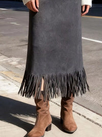 Person wearing a long dark gray suede skirt with fringe and brown boots on a street.