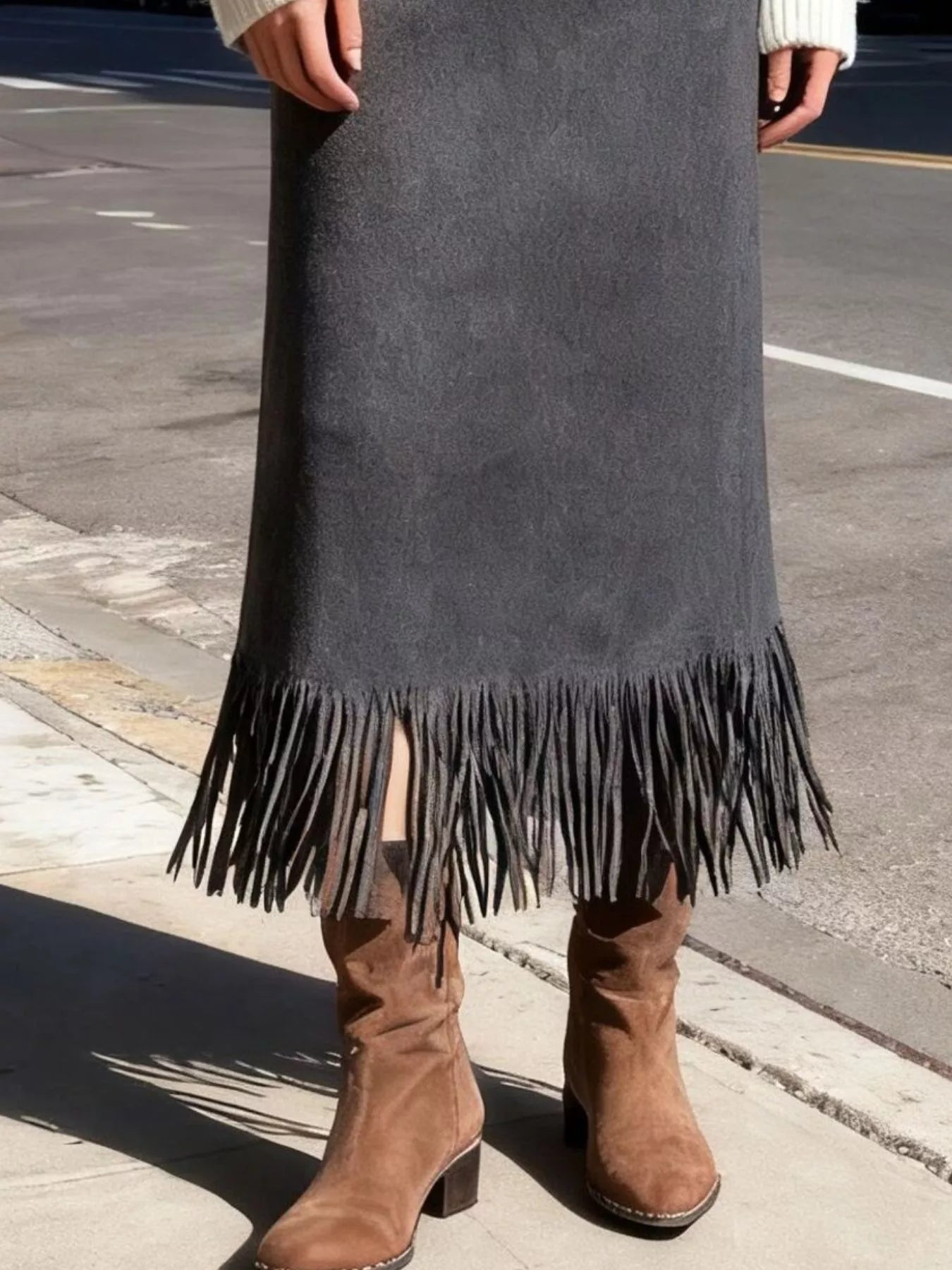 Person wearing a long dark gray suede skirt with fringe and brown boots on a street.