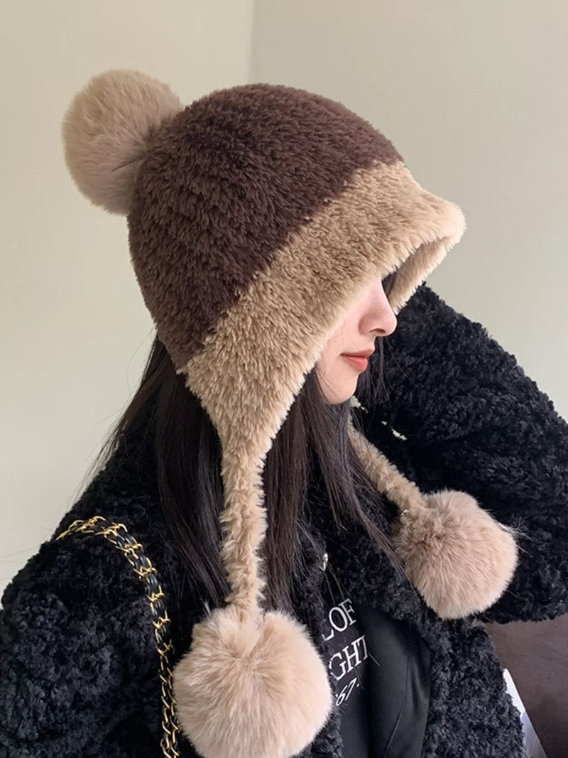 Person wearing a brown and beige fur-trimmed hat with pom-poms on a plain background