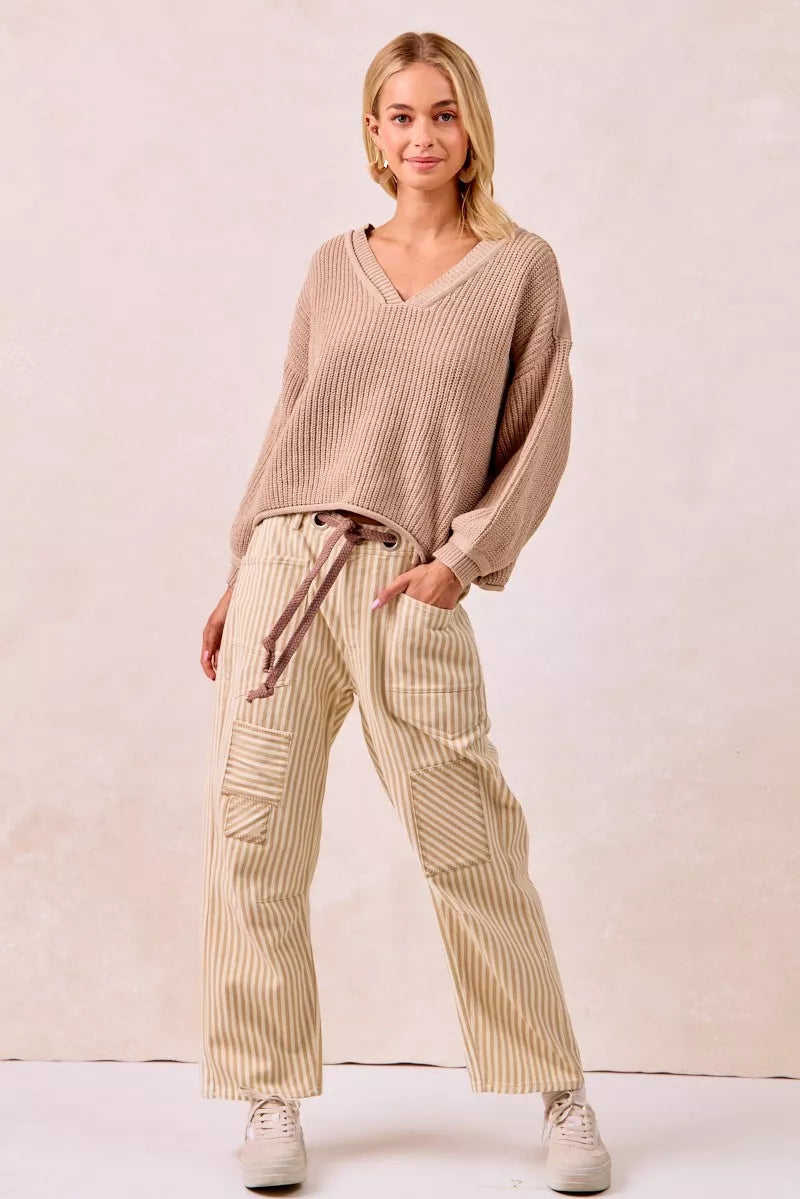 Woman wearing a beige knitted sweater and striped pants against a plain background