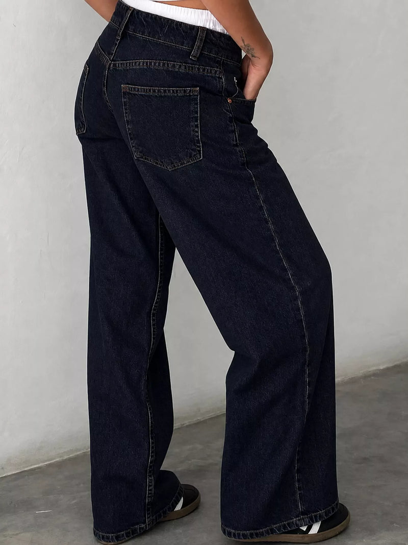 A person wearing dark navy wide-leg denim jeans, shown from the waist down, standing against a light gray wall.