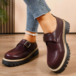 Brown leather shoes with buckles worn by a person on a wooden floor.