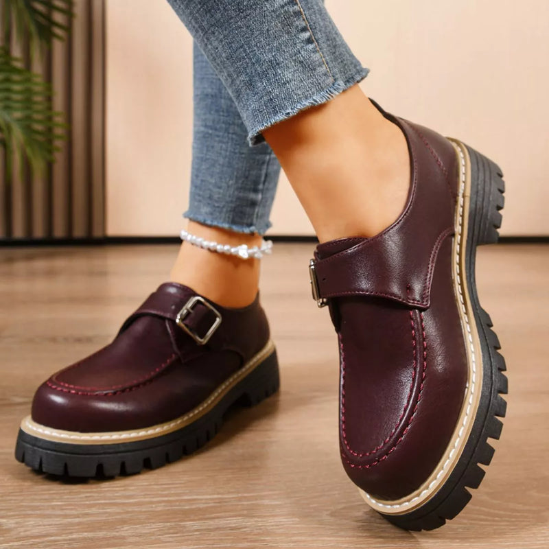 Brown leather shoes with buckles worn by a person on a wooden floor.