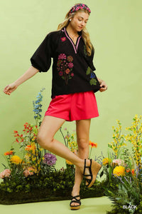 Woman in black floral embroidered top and red shorts standing in front of a floral background