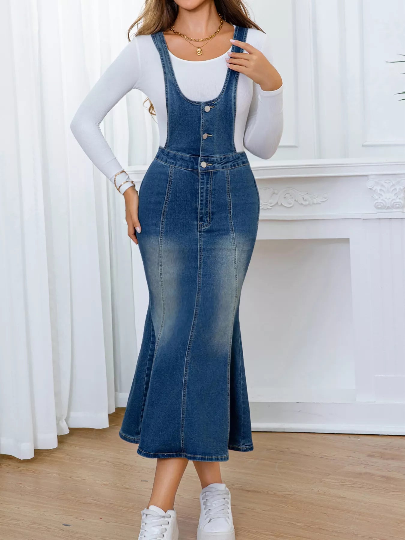 Person wearing a denim dress over a white long-sleeve shirt against a white curtain background