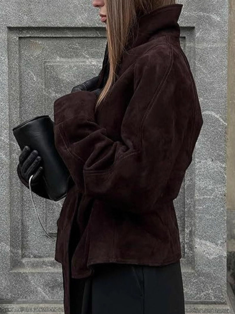 Person wearing a brown coat holding a black cup against a stone wall.