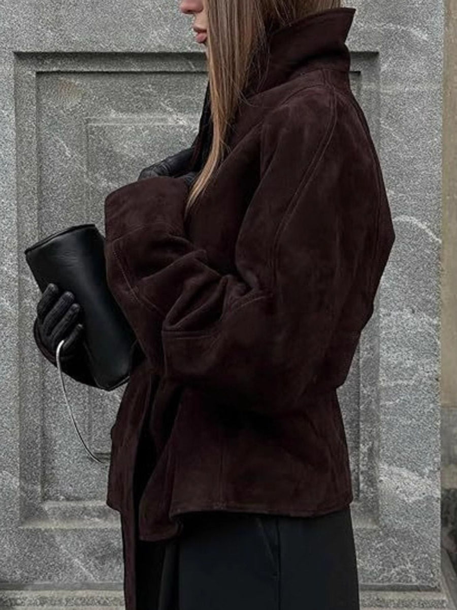 Person wearing a brown coat holding a black cup against a stone wall.
