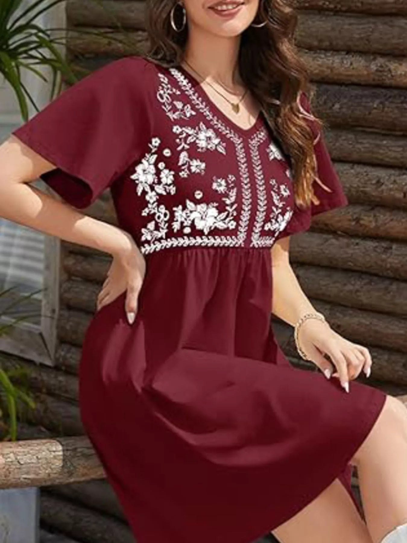 Woman wearing a burgundy dress with white floral embroidery against a wooden wall.