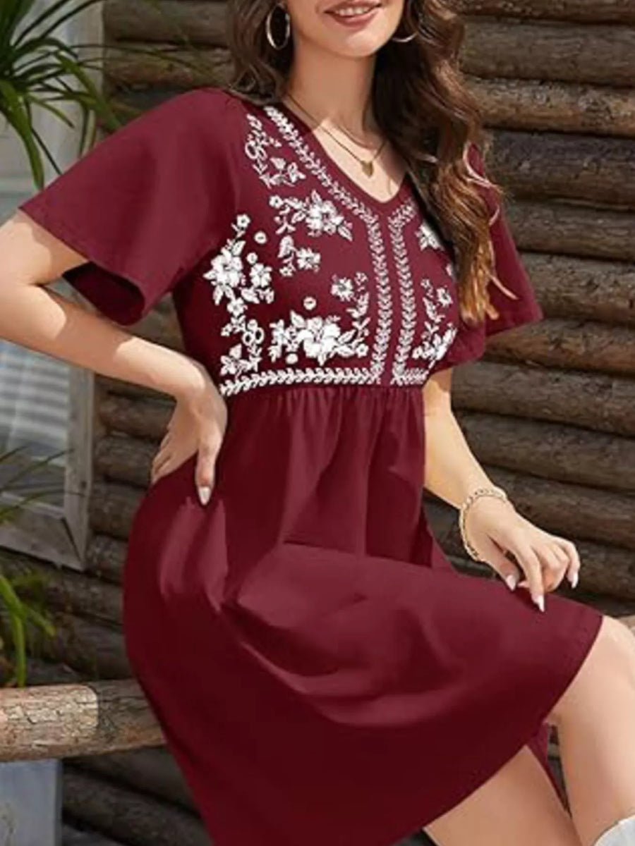 Woman wearing a burgundy dress with white floral embroidery against a wooden wall.