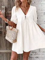 Woman wearing a white dress holding a woven bag against a rustic wooden and stone wall.