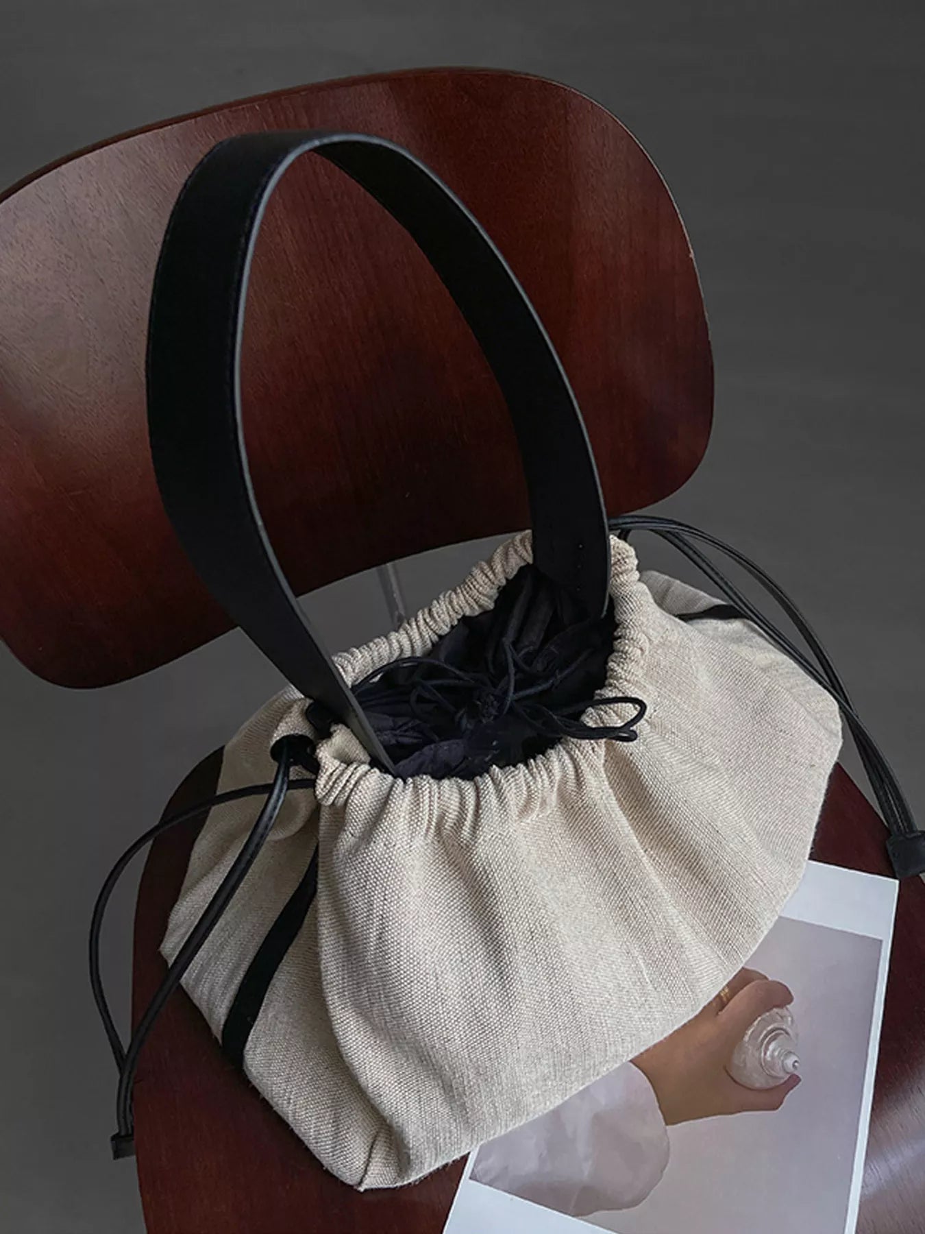 Beige fabric bag with black drawstring on a wooden chair, with a hand holding a bottle of water in the foreground.