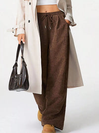 Person wearing brown wide-leg pants with a beige coat and holding a black handbag on a white background